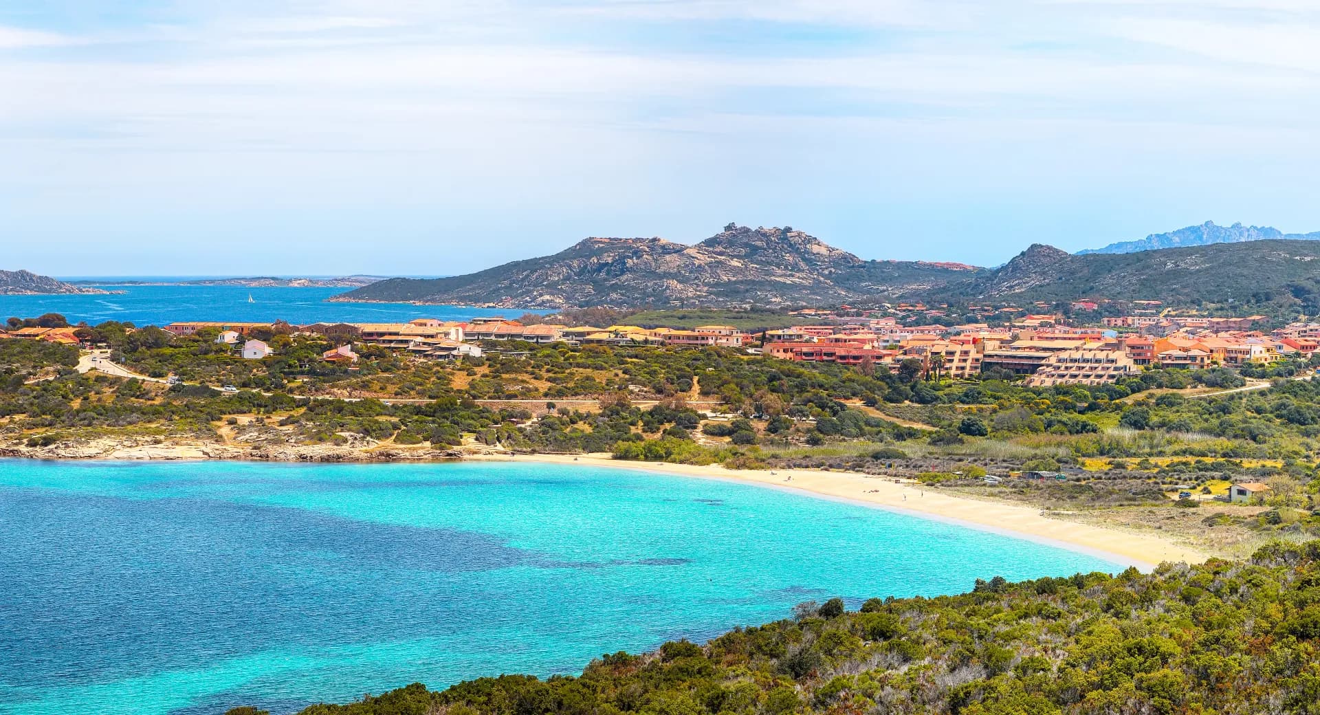Vue panoramique d'une baie avec une plage de sable fin, des eaux turquoise et un village côtier adossé à des collines verdoyantes sous un ciel clair