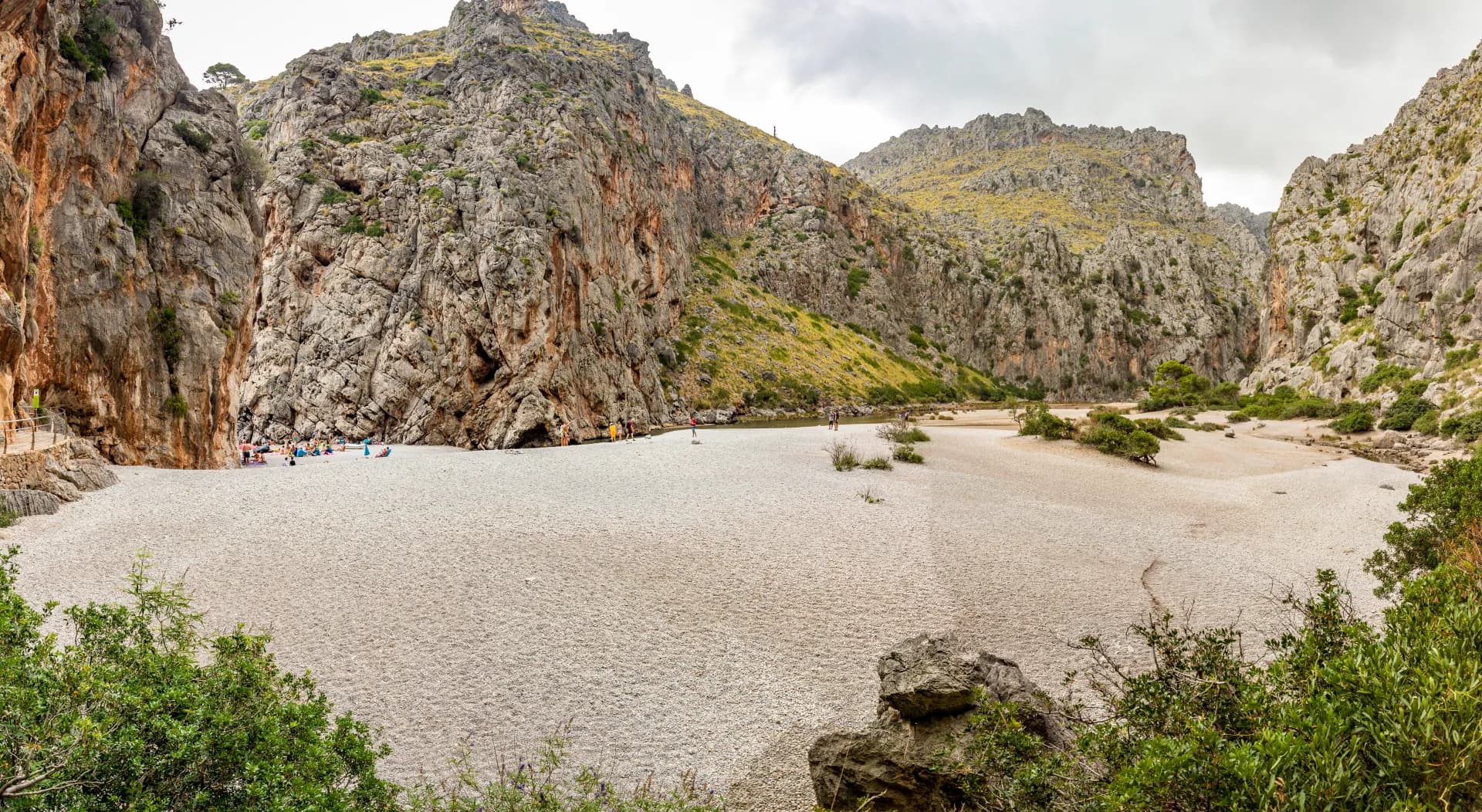 Vue panoramique d'une plage de galets entourée de hautes falaises rocheuses, avec des personnes se relaxant et un cours d'eau peu profond
