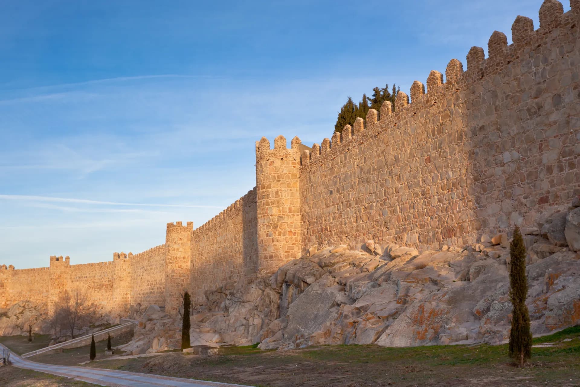Longue muraille médiévale en pierre avec des tours crénelées sous un ciel bleu clair, éclairée par le soleil couchant