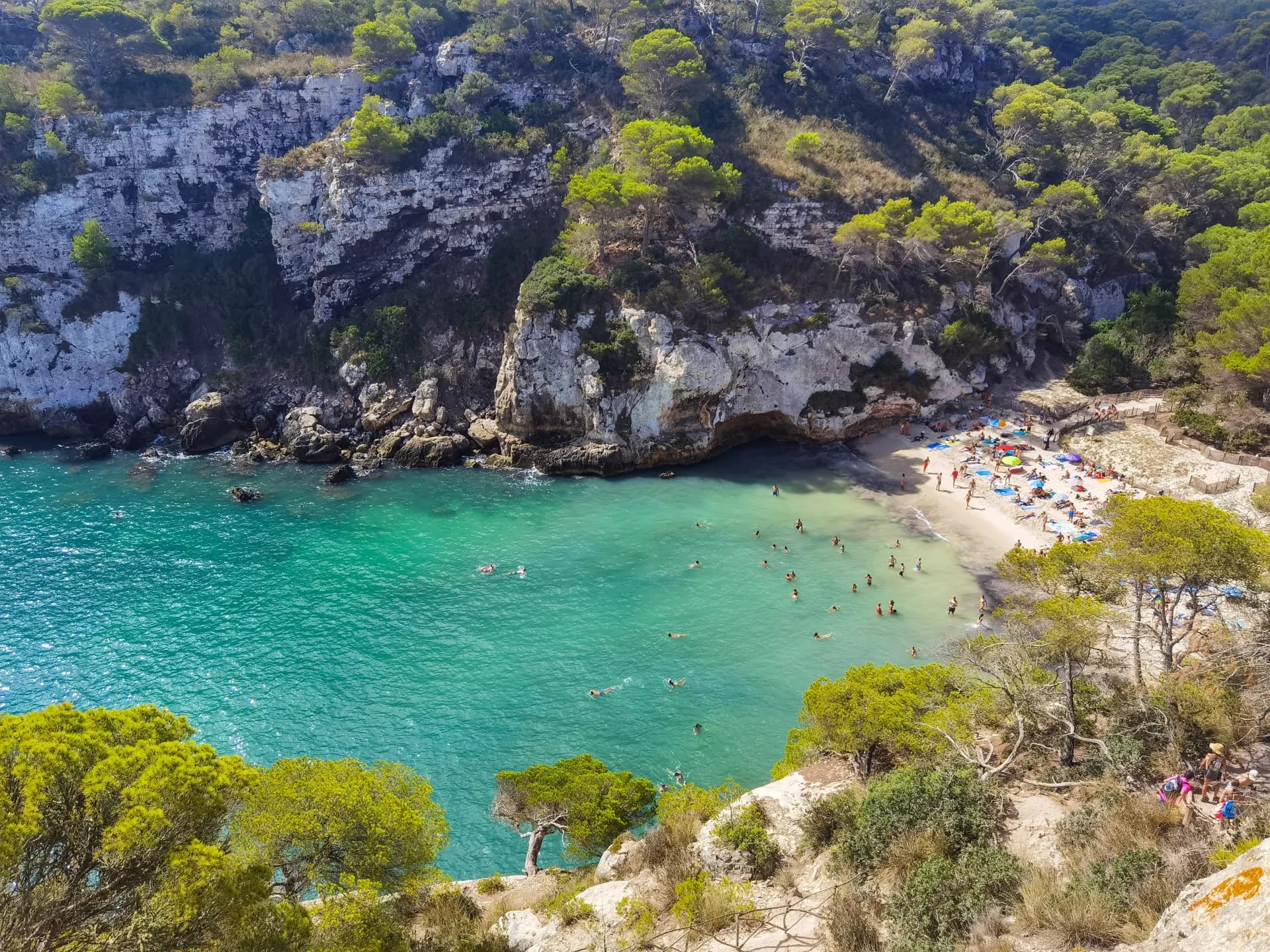 Vue aérienne d'une crique méditerranéenne avec une plage de sable fin, des eaux turquoise et des falaises rocheuses couvertes de végétation luxuriante