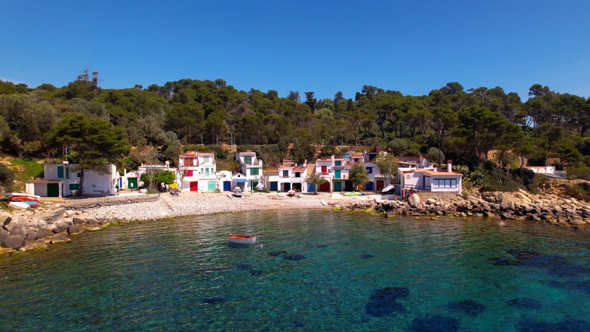 Vue aérienne d'un village de pêcheurs coloré au bord d'une crique rocheuse, avec des eaux turquoise claires et une forêt méditerranéenne luxuriante en arrière-plan sous un ciel bleu.