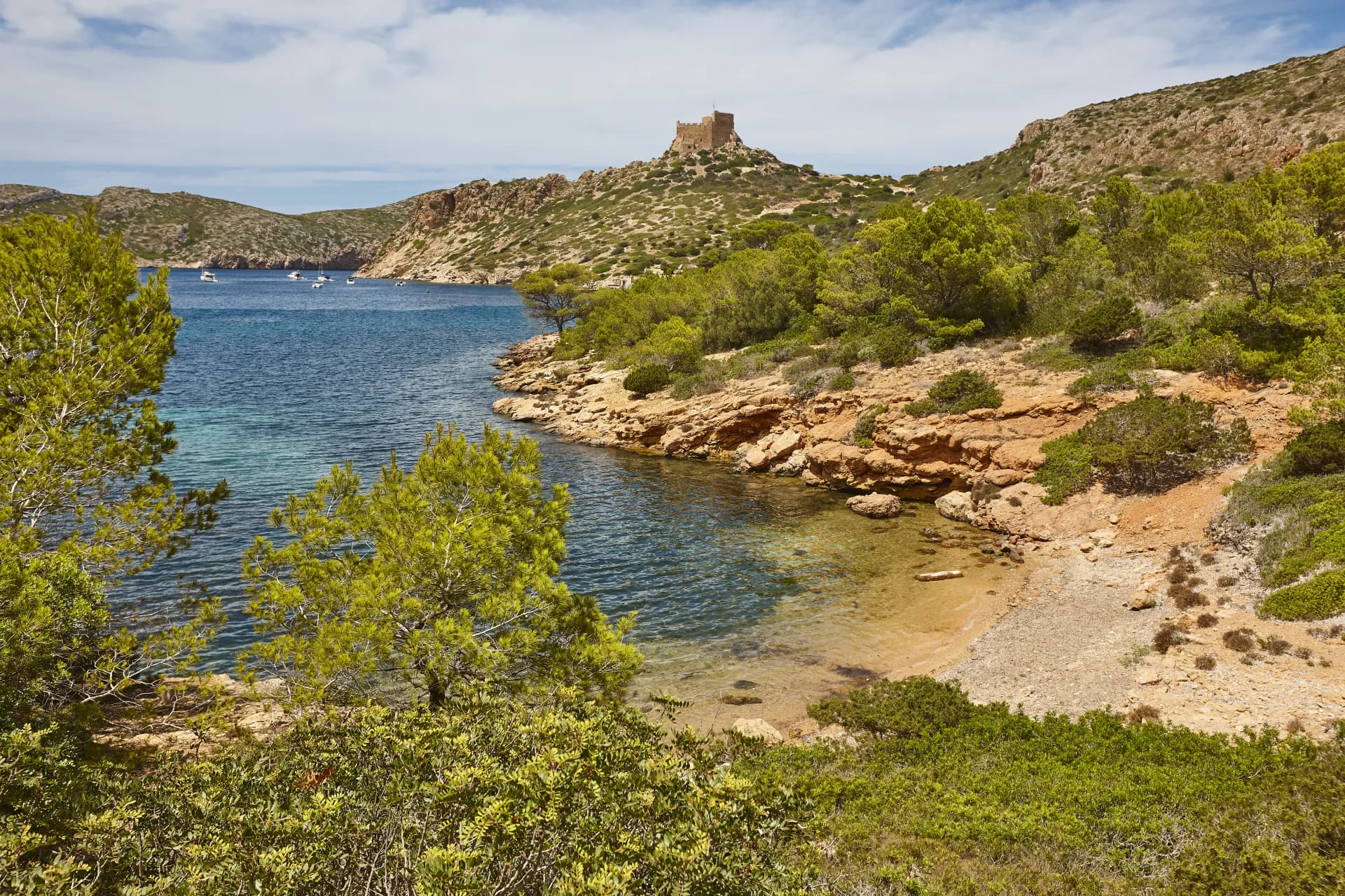 Vue panoramique d'une crique rocheuse avec des eaux turquoise, bordée de pins verts et de végétation méditerranéenne sous un ciel nuageux. Un ancien bâtiment en pierre est visible sur une colline au loin.