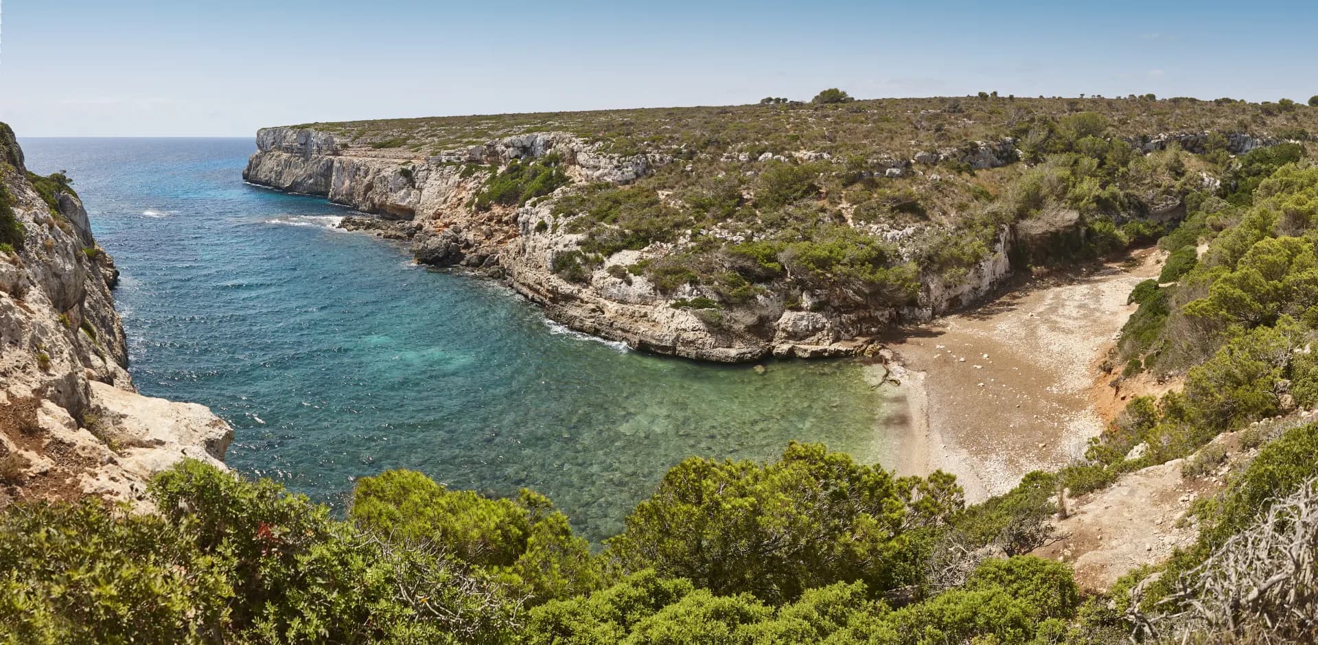 Vue panoramique d'une crique rocheuse avec des eaux turquoise et une petite plage de galets, entourée de végétation méditerranéenne sous un ciel bleu clair.