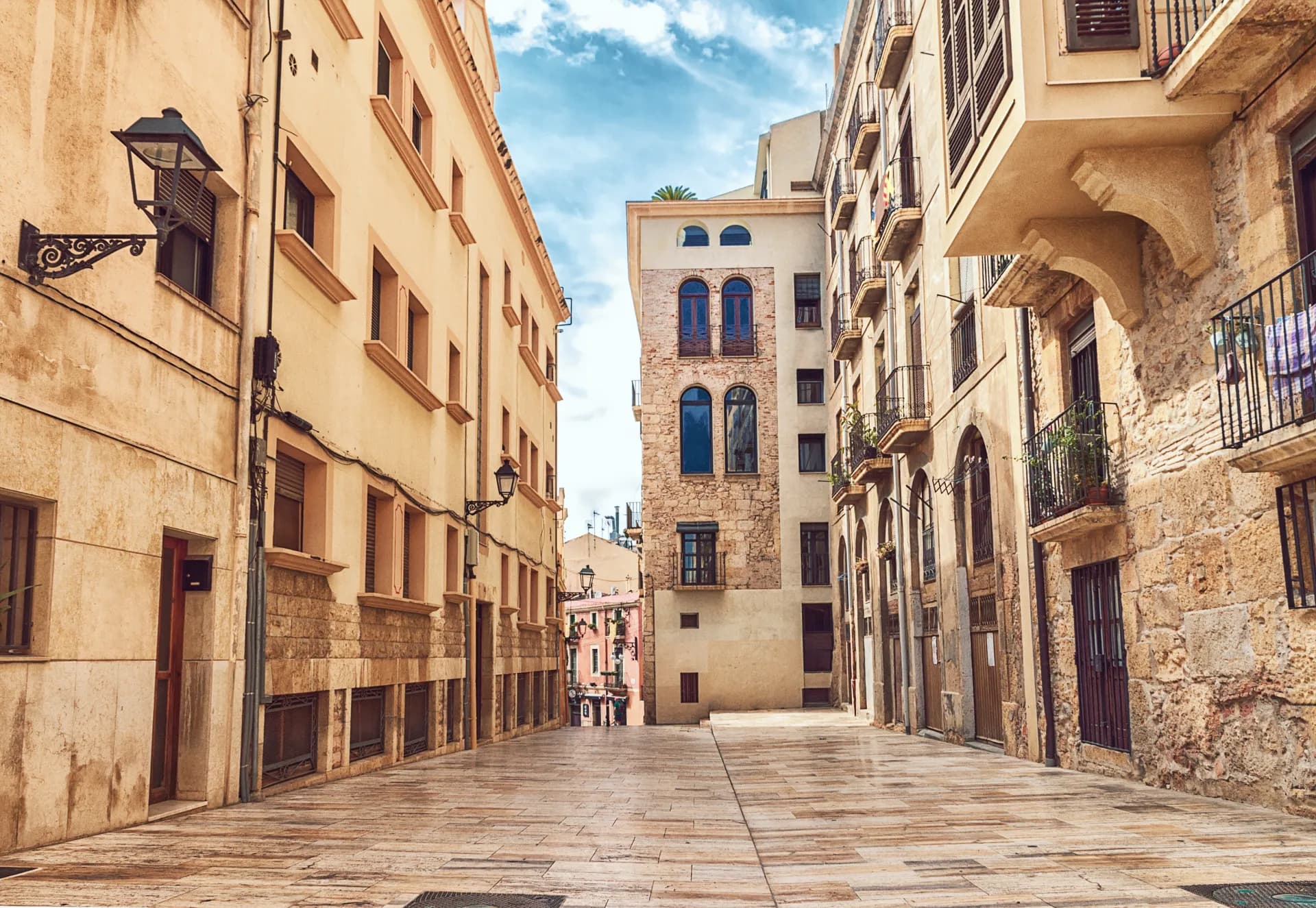Rue étroite et vide dans une vieille ville méditerranéenne, avec des bâtiments aux façades ocres et des balcons en fer forgé sous un ciel bleu clair.