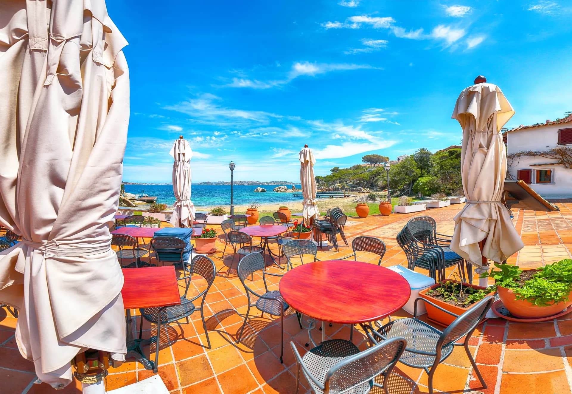 Terrasse de restaurant en bord de mer avec tables et chaises, parasols repliés, plage de sable et mer turquoise sous un ciel bleu clair