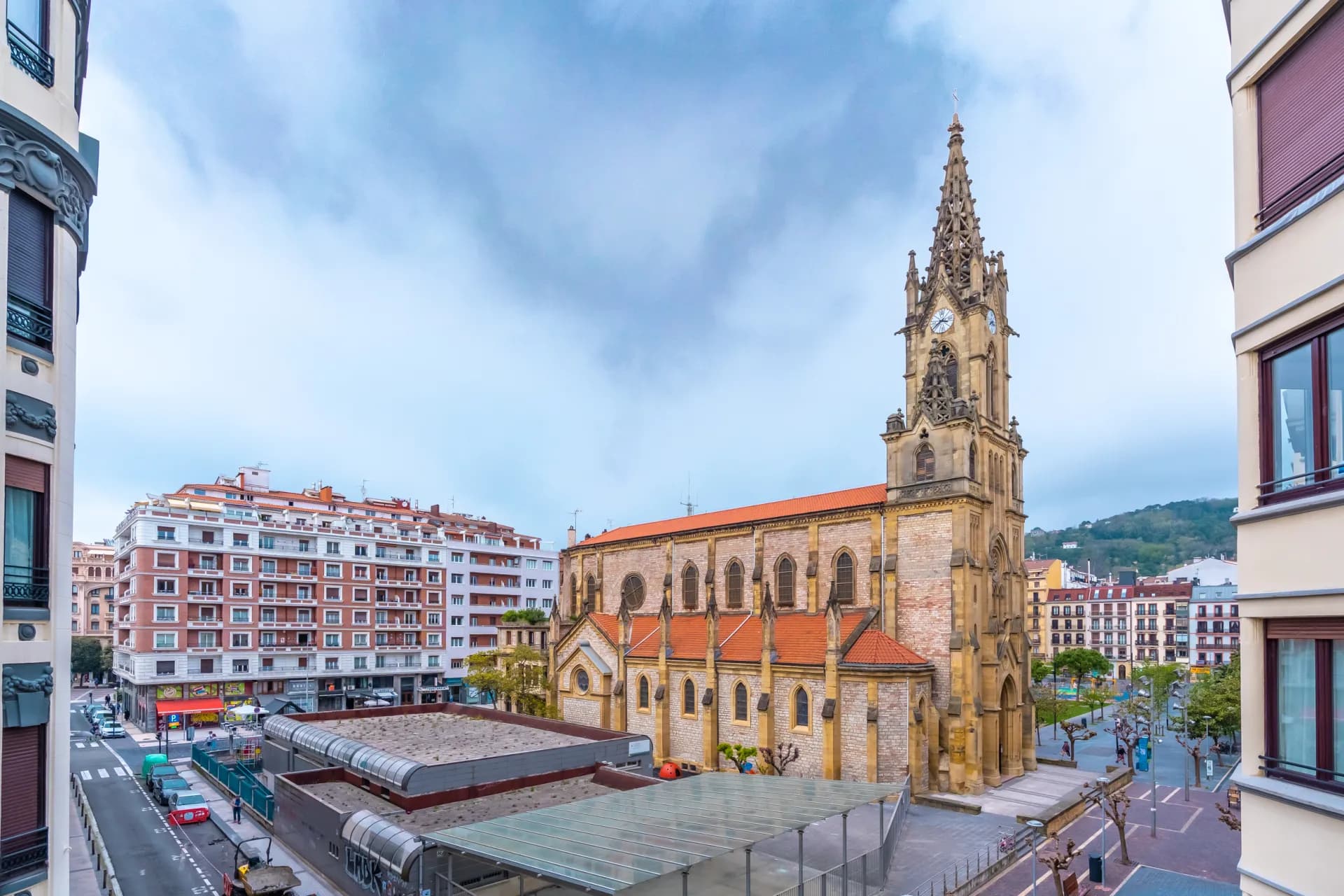 Vue d'une église ancienne avec un clocher imposant et des toits en tuiles rouges, entourée de bâtiments urbains modernes sous un ciel nuageux.