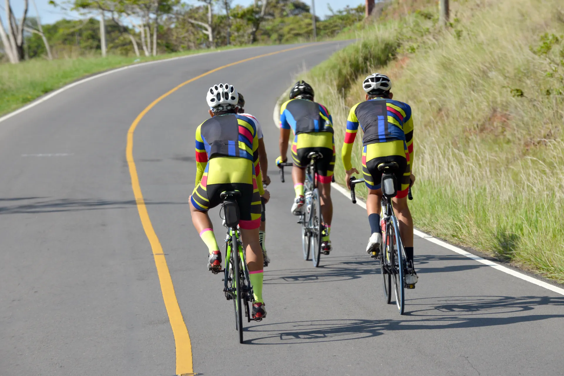 Trois cyclistes en tenue colorée montent une route goudronnée sinueuse bordée d'herbe haute sous un ciel clair