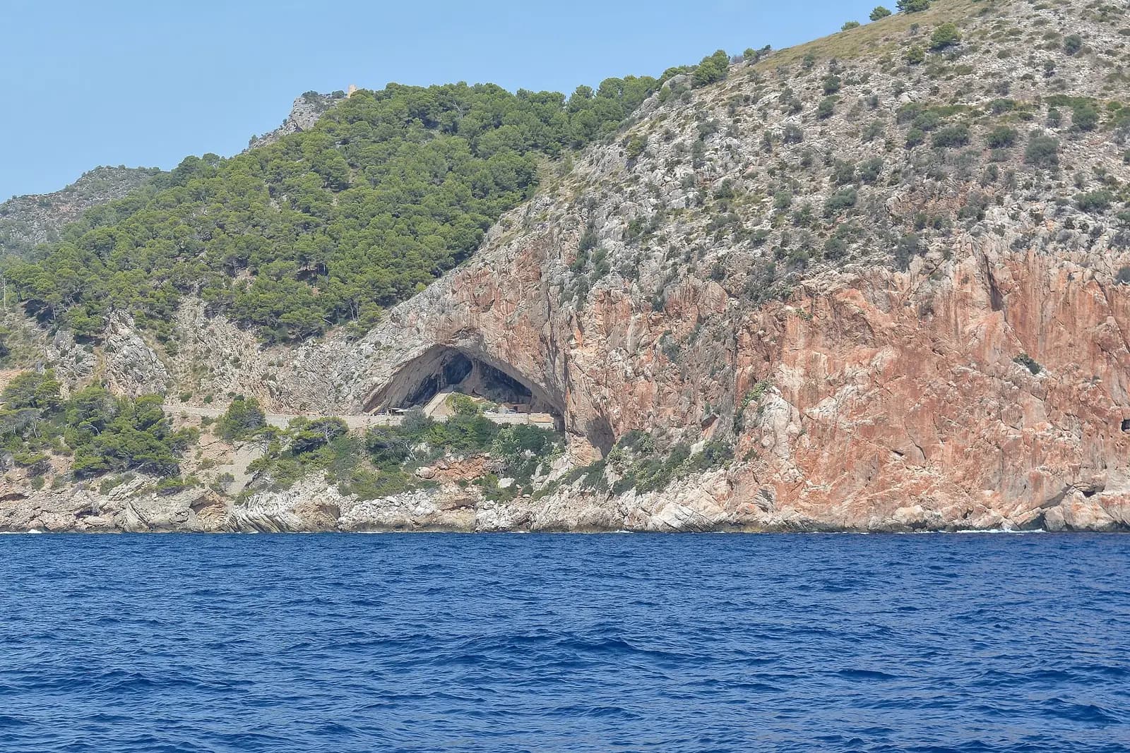 Falaise rouge et grise du Cap Vermell vue depuis la mer, avec la grande ouverture en arche des grottes d’Artà au pied de la pente